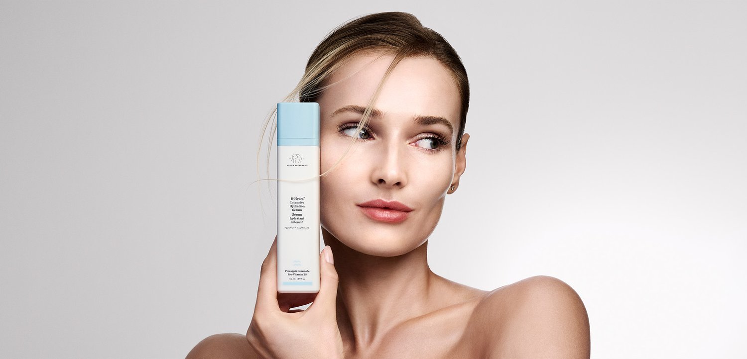 Frau mit heller Haut und blondem Haar hält ein blau-weißes Hautpflegeprodukt dicht an ihr Gesicht vor einem schlichten, hellen Hintergrund.