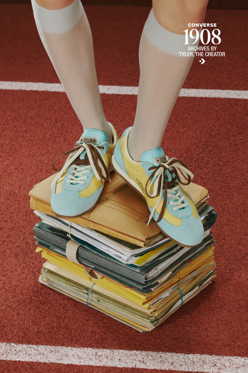 Piernas con calcetines blancos y zapatillas Converse azul y amarillo sobre archivos y carpetas vintage apilados sobre una pista roja.