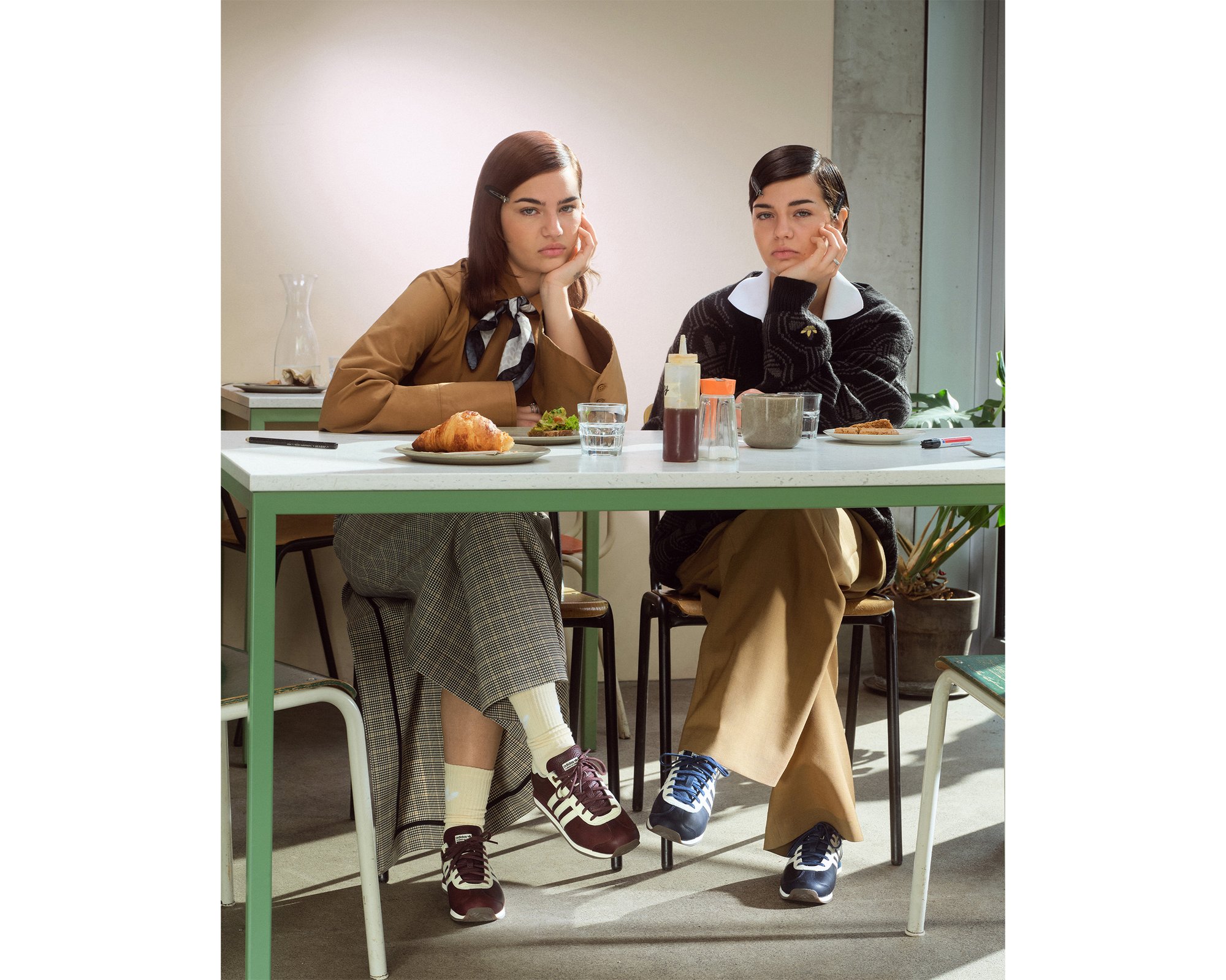Twee vrouwen zitten aan een tafel in een café, met nadenkende gezichten. Eten en drinken zijn op de tafel gerangschikt, met planten op de achtergrond.