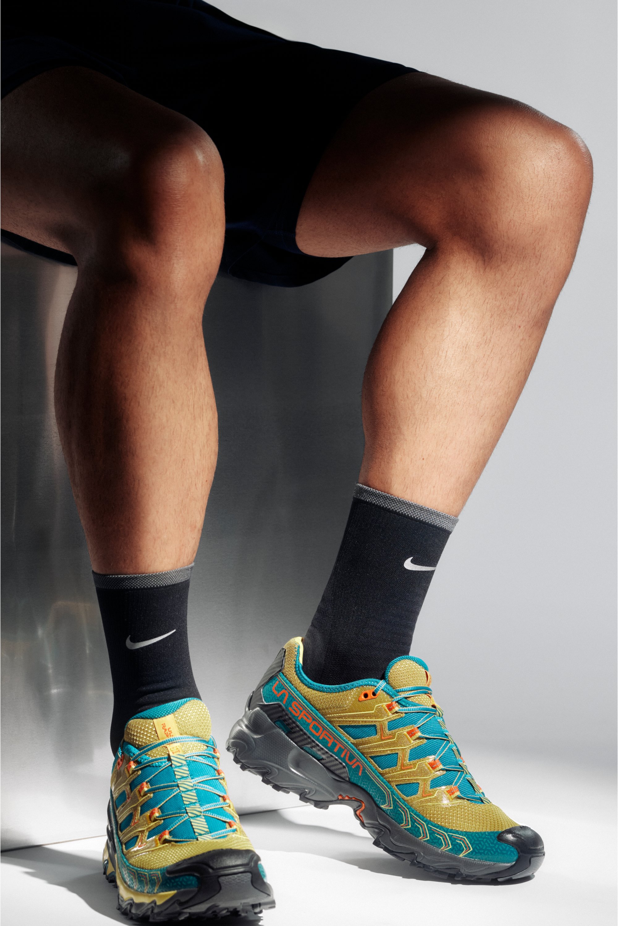 Athletic legs in black shorts wear bright yellow La Sportiva shoes with turquoise accents and black Nike socks against a neutral background.