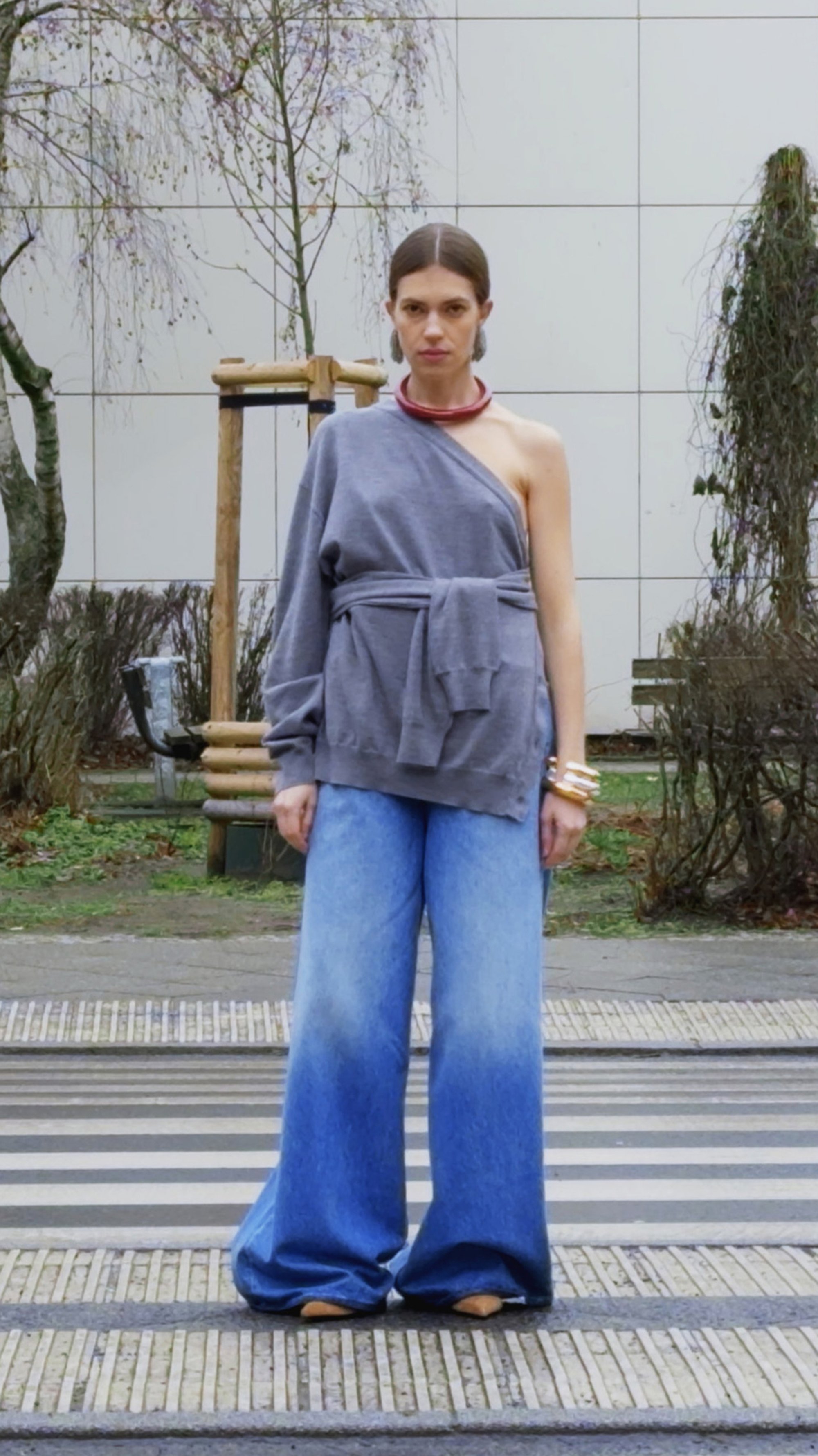 Una mujer se stands con confianza en una blusa gris de un solo hombro y pantalones de mezclilla de pierna ancha en una calle con árboles y un edificio al fondo.
