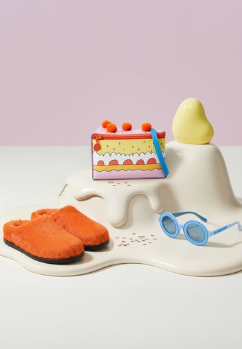 Leuchtend orangefarbene, flauschige Hausschuhe, bunte kleine Tasche mit Bommel, blaue runde Sonnenbrille und eine gelbe abstrakte Skulptur auf einer welligen cremefarbenen Oberfläche.