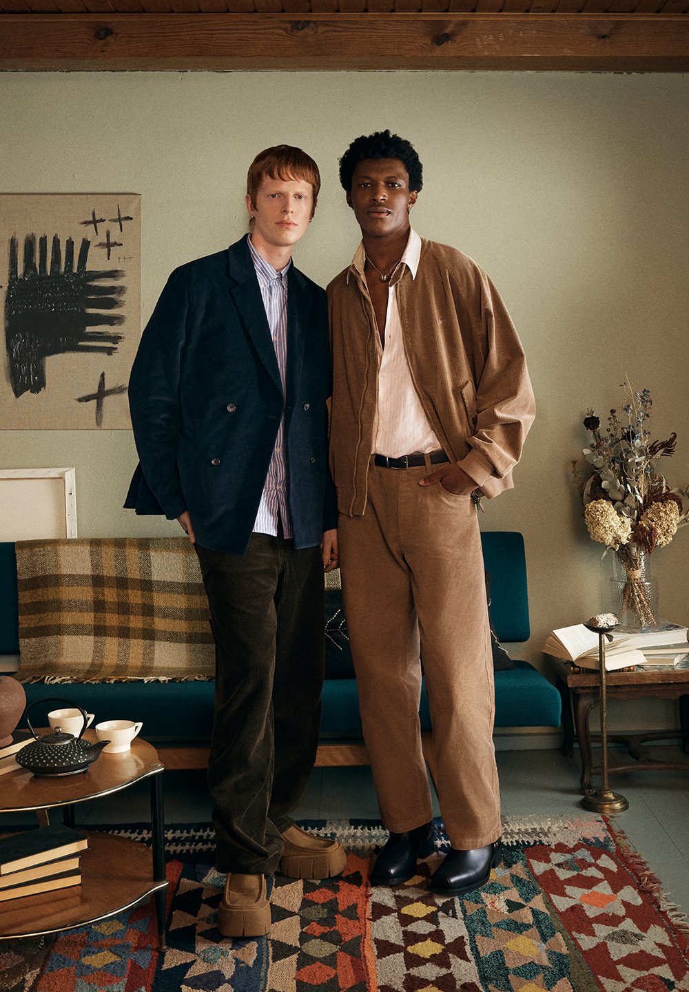 Két személy áll együtt egy szobában. Az egyik tengerészkék, kétsoros kabátot visel csíkos ing felett; a másik barna bombázókabátot hord.