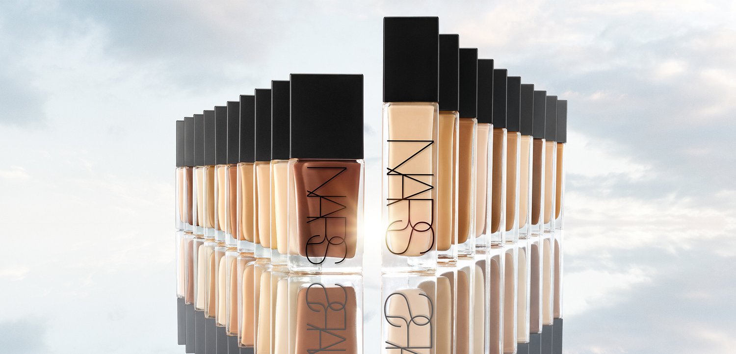 File di flaconi di fondotinta in tonalità sfumate dal più scuro al più chiaro con tappi neri e logo del marchio, riflessi su una superficie lucida.