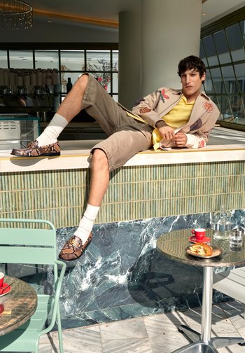 Man in patterned jacket, yellow shirt, and shorts reclining on a café counter with a croissant, red cup, and water bottle on a nearby table.
