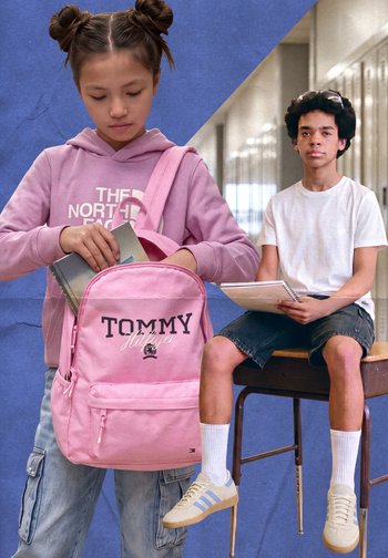 Ragazza con felpa rosa con cappuccio che mette un libro in uno zaino rosa Tommy Hilfiger, ragazzo con t-shirt bianca e pantaloncini seduto a una scrivania che tiene un quaderno.