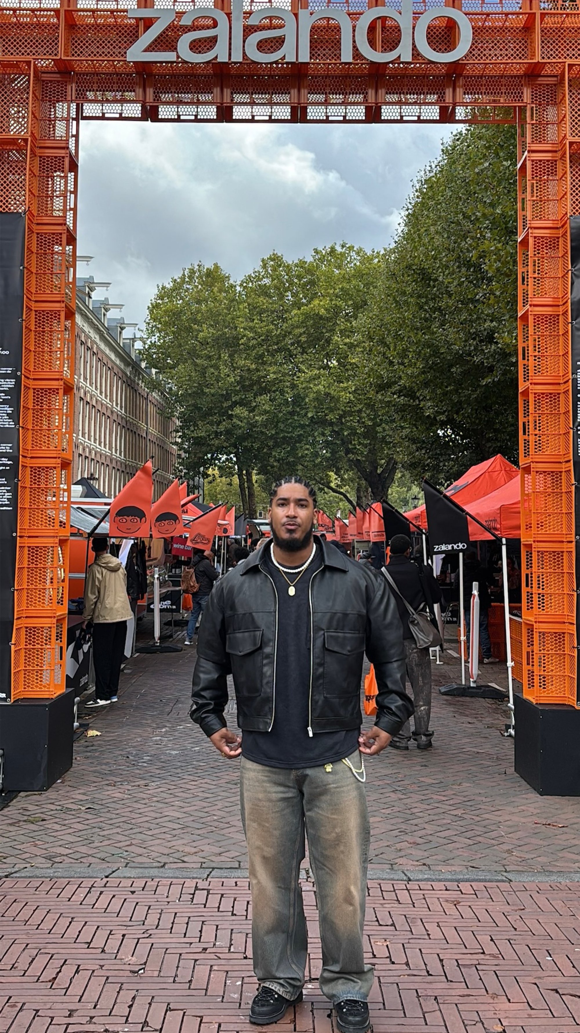 Man in een zwarte leren jas en vervaagde jeans staat onder een oranje Zalando-bogen bij een buitenmarkt met tenten en vlaggen.