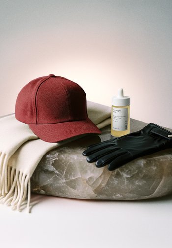 Czerwona czapka baseballowa na kremowym szaliku, czarna skórzana rękawica oraz mała bursztynowa buteleczka zakraplacza na okrągłej marmurowej powierzchni.