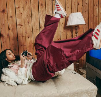 Eine Person mit langen Haaren entspannt sich auf einem Sofa und hält einen Spielcontroller in der Hand. Sie trägt einen weißen Pullover und große bordeauxfarbene Hosen, mit hochgelegten Füßen.
