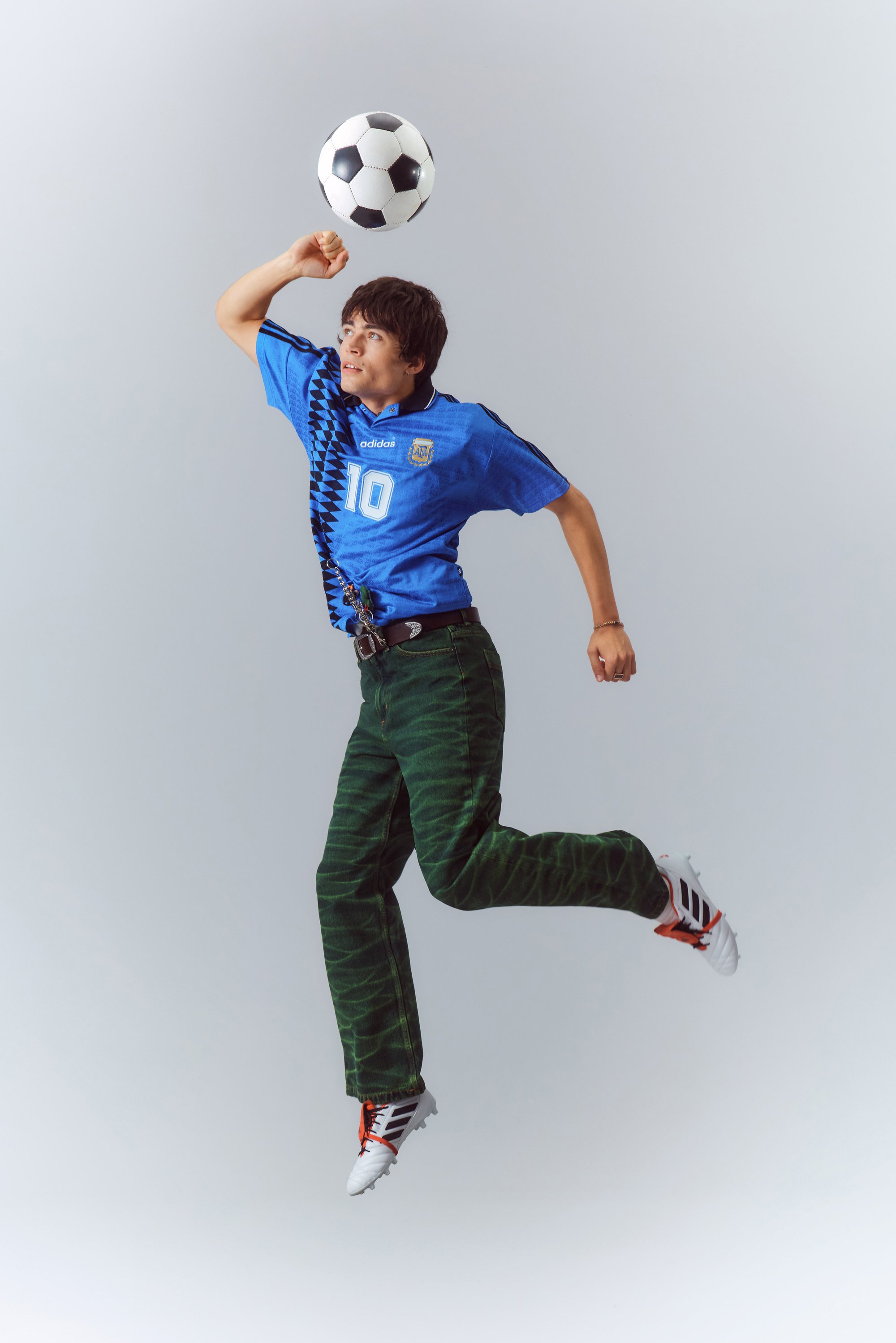 A young athlete in a blue soccer jersey jumps while preparing to kick a black and white soccer ball. He wears green pants and white cleats.