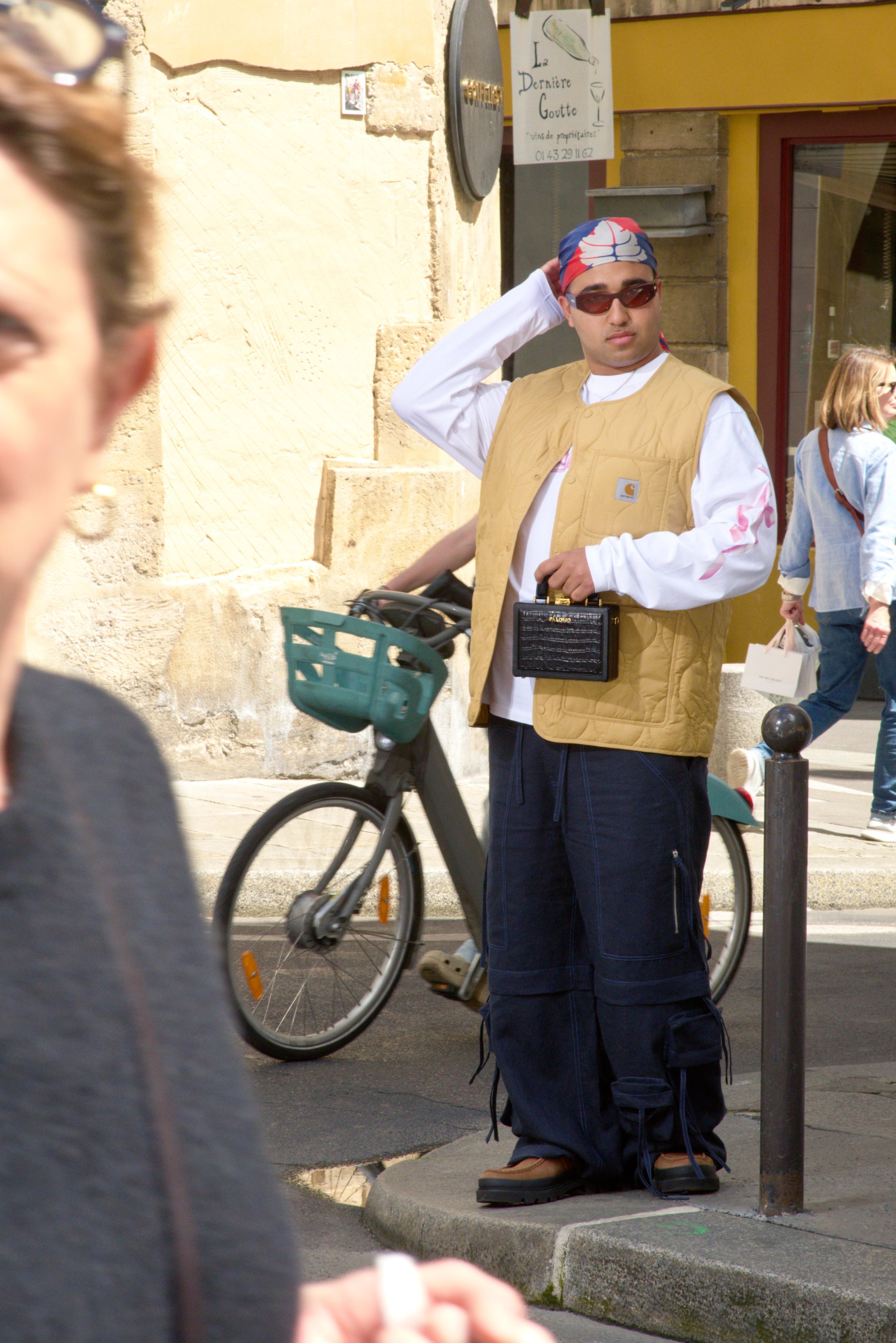 Una persona con un chaleco amarillo y pantalones anchos azules está de pie junto a una bicicleta, ajustándose una cinta para la cabeza. La gente pasa en una escena callejera animada.
