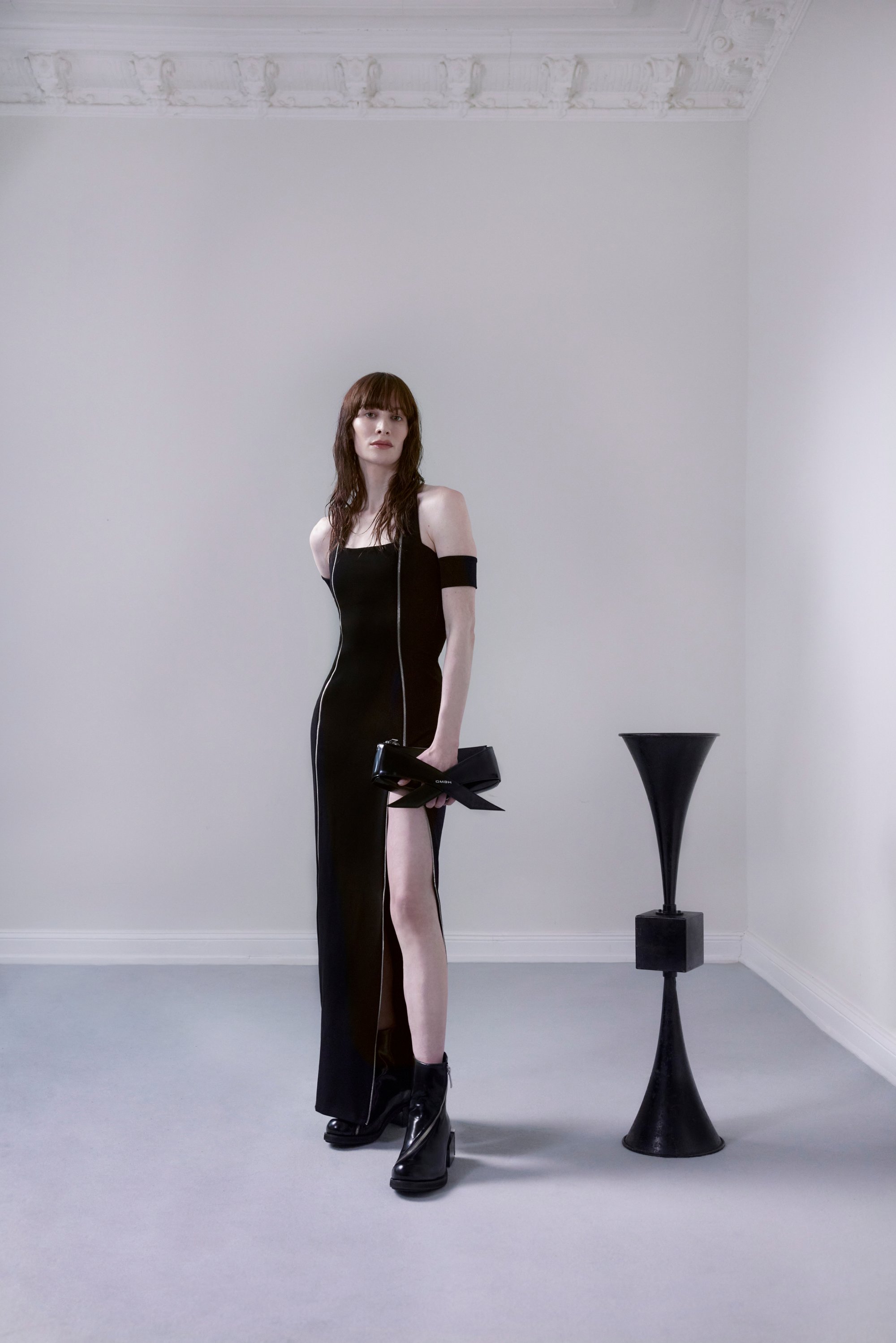 Una modella in un elegante abito nero posa con sicurezza, tenendo una borsa nera, su uno sfondo minimalista grigio chiaro con una scultura moderna.