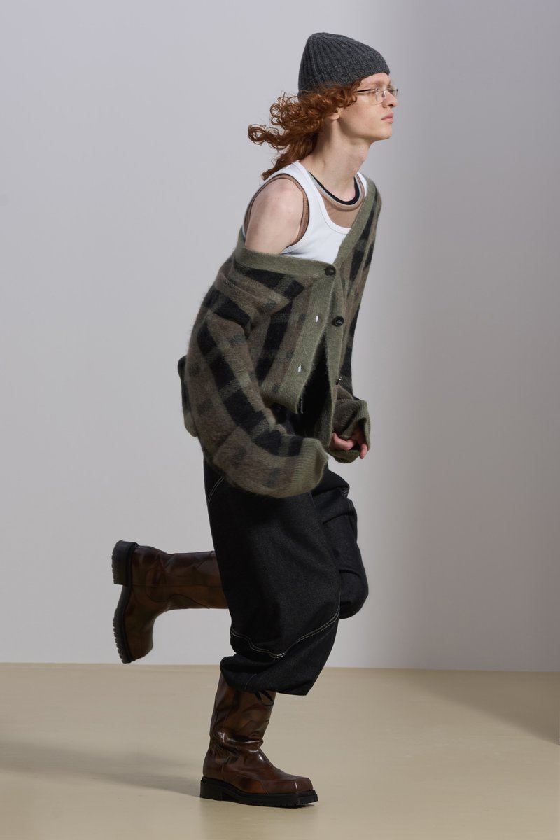 Una persona con cabello rizado, que lleva un gorro gris, un cárdigan oversize, una camiseta sin mangas y pantalones negros, camina con confianza sobre un suelo beige.