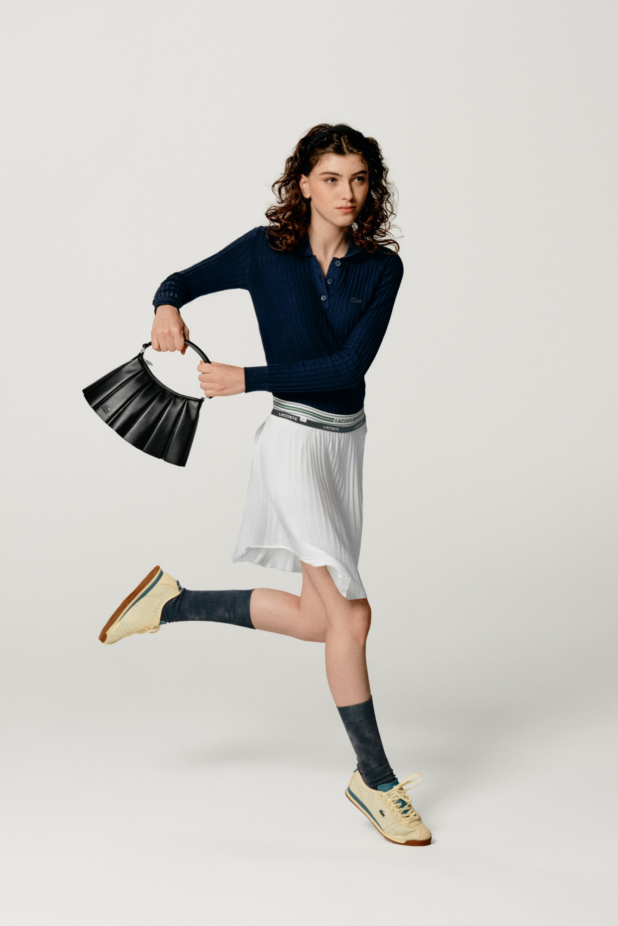 Giovane donna con capelli ricci che corre, indossando una maglietta blu marina, una gonna bianca e sneakers, mentre tiene una borsa nera, su uno sfondo chiaro.