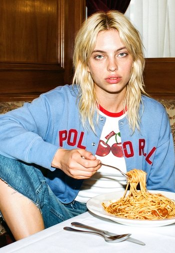 Jeune personne aux cheveux blonds portant une veste bleue et un jean, mangeant des spaghettis à une table avec des ustensiles sur une nappe blanche.