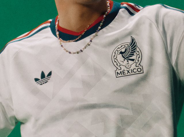 Persoon draagt een wit Mexico voetbalshirt met een rode en groene kraag, Adidas-logo op de linkerborst en het Mexicaanse embleem op de rechterborst.