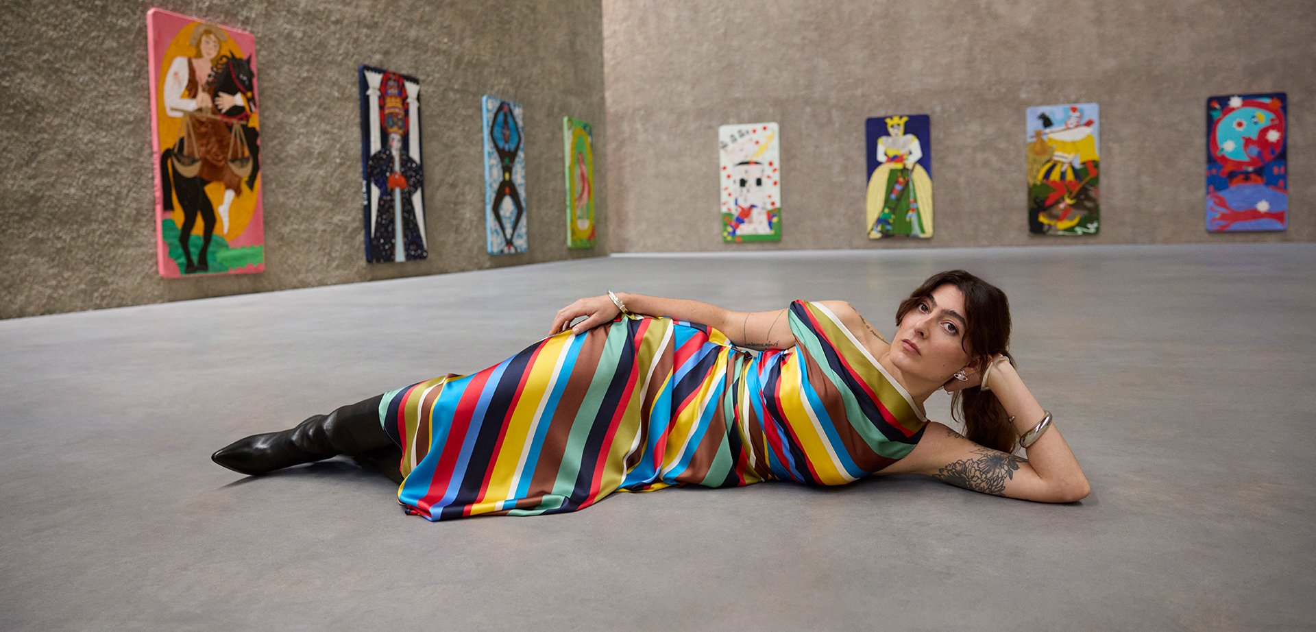 Multicoloured striped silk dress with wide horizontal stripes in red, blue, green, and brown; person reclining on a smooth floor in an art space.
