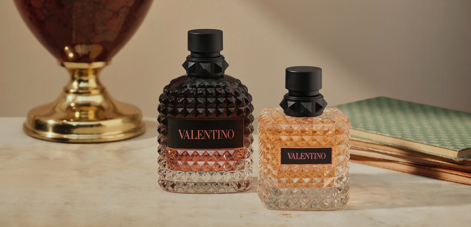 Twee vierkante Valentino parfumflesjes met zwarte doppen staan op een marmeren oppervlak naast een gouden vaas en gestapelde groene boeken.
