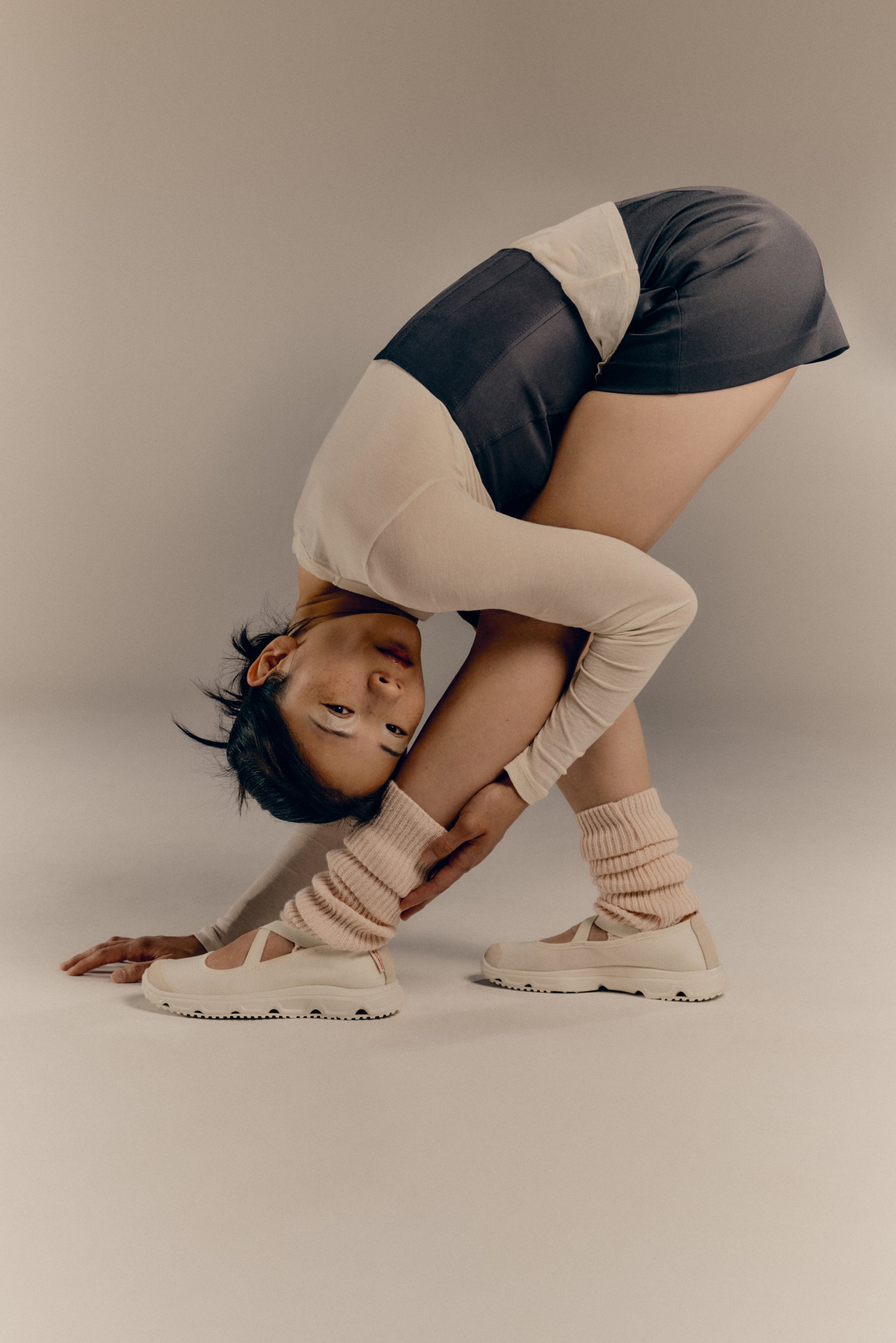 A person performs a deep stretch, bending forward with one arm holding a leg, wearing a beige top, shorts, and pink leg warmers.