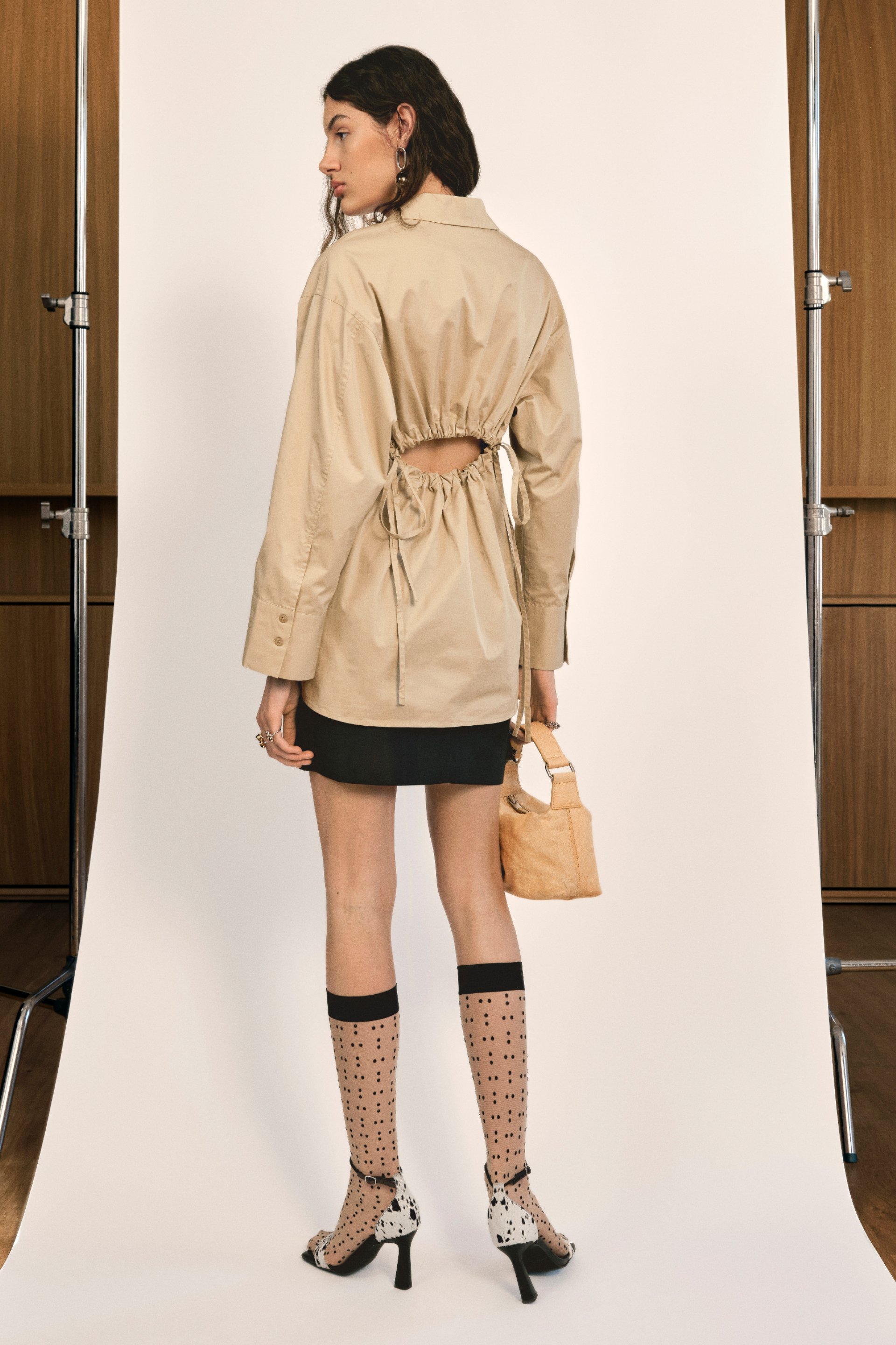 La modelo está de espaldas a la cámara, vistiendo una camisa beige con la espalda descubierta, una falda negra, calcetas de lunares y tacones con manchas, sosteniendo un pequeño bolso beige.