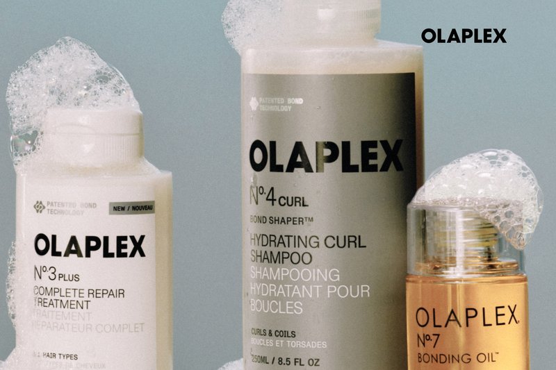 Three Olaplex hair care bottles with foam on top, labeled No. 3 Plus Complete Repair Treatment, No. 4 Curl Hydrating Shampoo, and No. 7 Bonding Oil.
