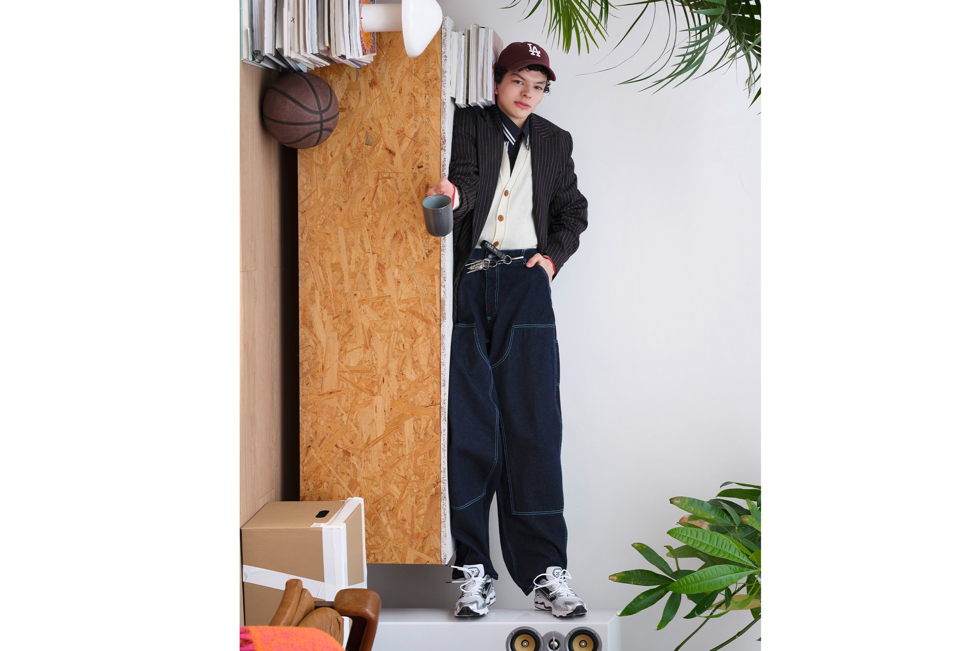 A young person stands in a stylish outfit with oversized denim pants, holding a mug, surrounded by books, a basketball, and plants.