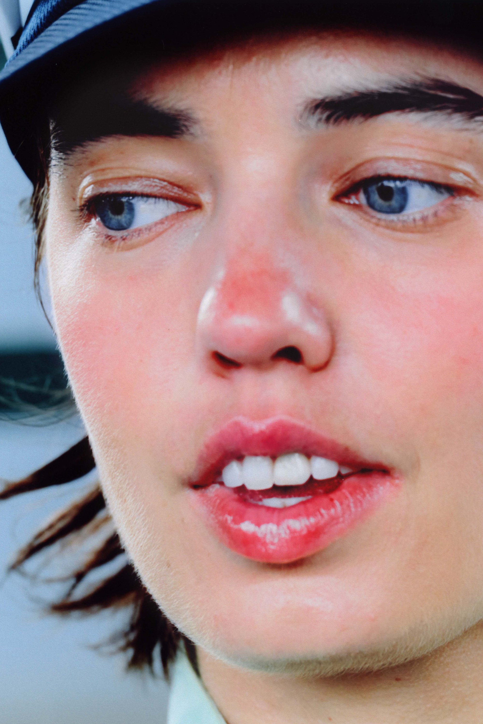 Close-up of a face with smooth skin, light pink lips, and blue eyes. Hair is dark and wavy, partially visible at the sides.