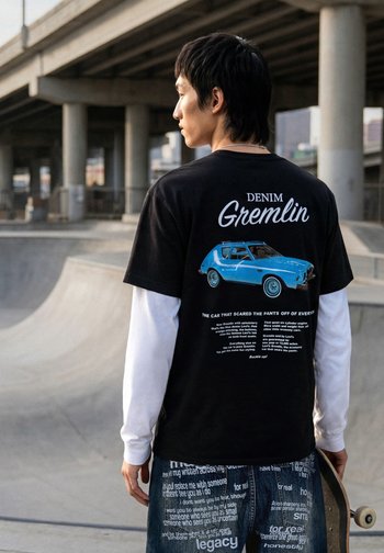 Jeune homme tenant un skateboard sous un passage surélevé, portant un t-shirt noir avec un graphisme de voiture bleue et du texte blanc au dos.