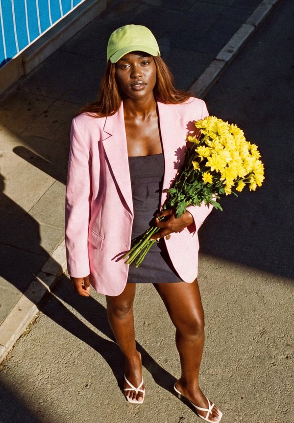 Femme debout dans une rue ensoleillée tenant des fleurs jaunes, portant un blazer rose, une robe noire, une casquette vert lime et des sandales blanches.