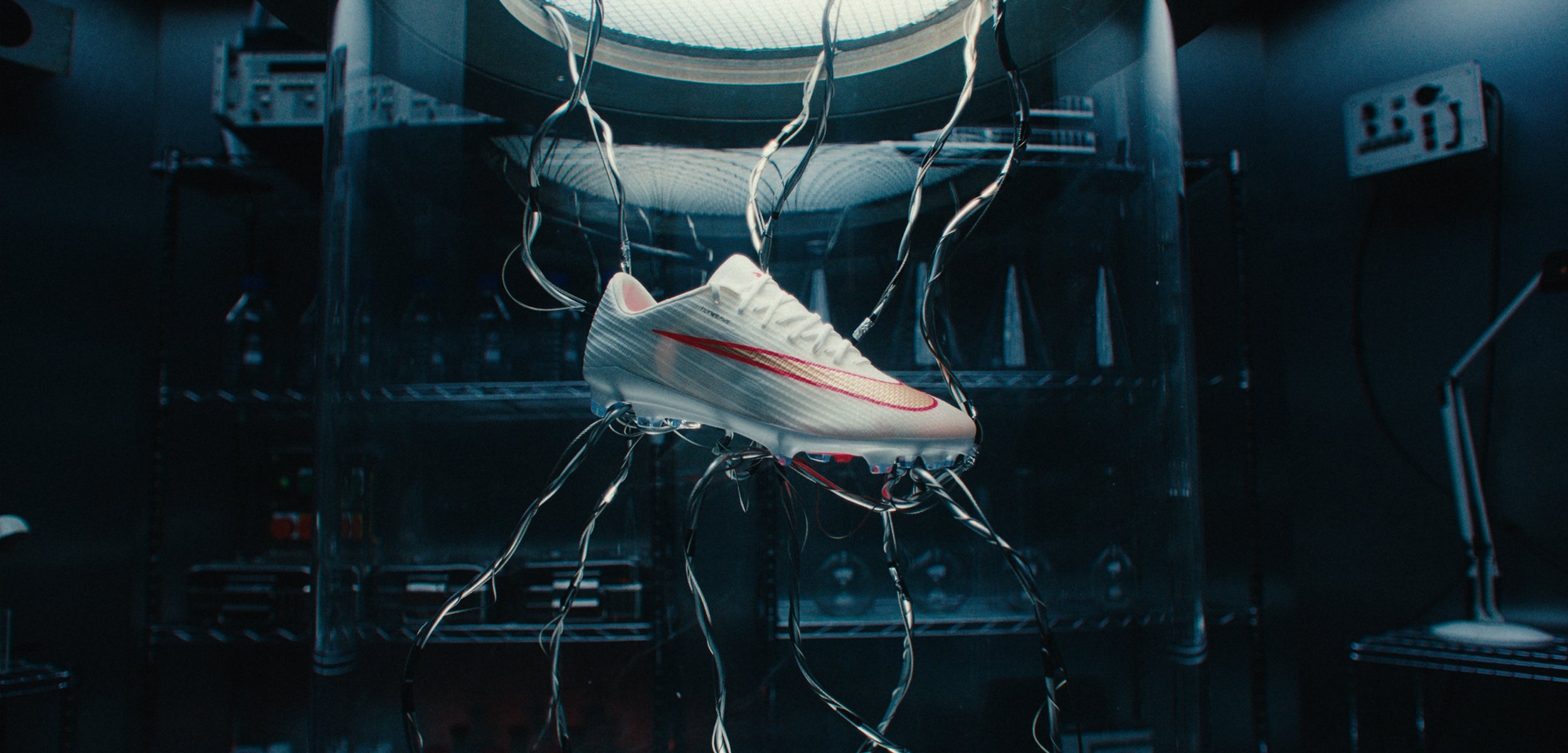 Weißer Sportschuh mit rotem Swoosh, an mehreren dünnen schwarzen Drähten hängend, in einem dunklen Laborumfeld mit hellem Licht von oben.