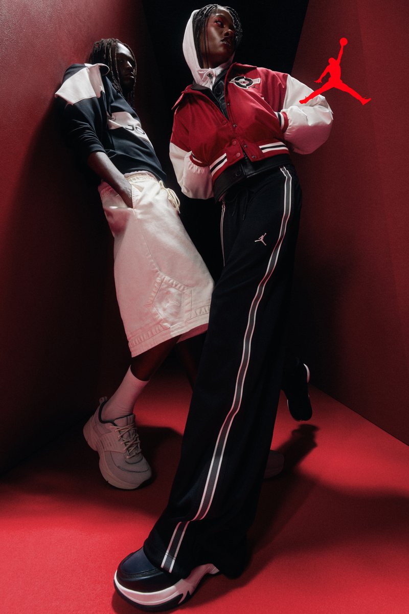Dos personas posan en una esquina iluminada en rojo vistiendo ropa deportiva de la marca Jordan, que incluye sudadera con capucha, chaqueta, pantalones cortos y pantalones de chándal con zapatillas.