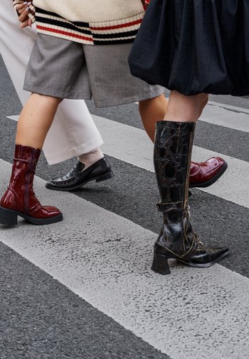 Three pairs of footwear: a burgundy leather ankle boot, a black textured boot with a pointed toe, and a black loafer, crossing a street.