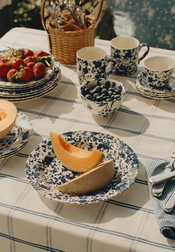 Μπλε και λευκά πιάτα, φλιτζάνια και μπολ με βατόμουρα, φέτες πεπόνι και φράουλες πάνω σε λευκό τραπεζομάντηλο με μπλε ρίγες.