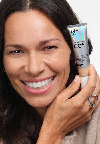 Uśmiechnięta kobieta z długimi ciemnymi włosami trzyma przy twarzy tubkę kremu It Cosmetics CC+ z filtrem SPF 50+.