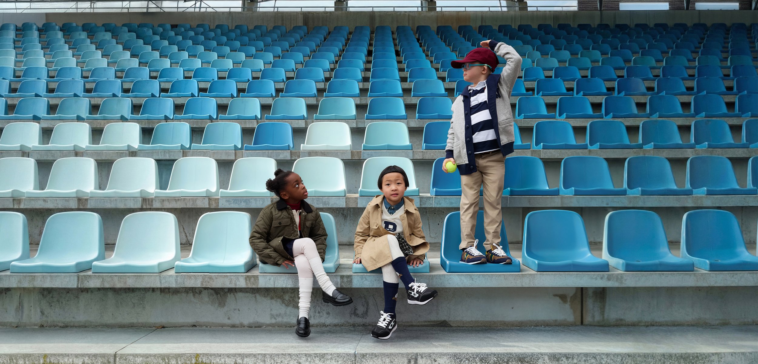 Drei Kinder in Freizeitkleidung sitzen auf blauen Tribünenplätzen. Das Mädchen in einer grünen Jacke und weißen Strumpfhosen blickt nach oben, während ein Junge über ihr steht und einen Tennisball hält.