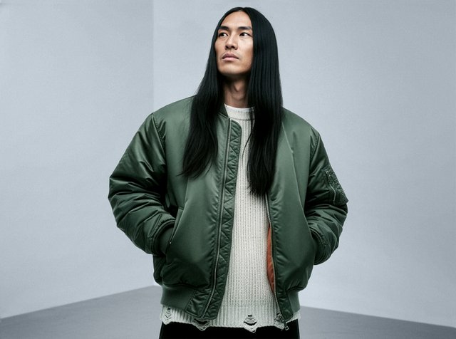 Olivgrön bomberjacka med satinfinish, ribbade muddar och en dragkedjeförsedd ficka på ärmen, bärs över en krämfärgad stickad tröja.