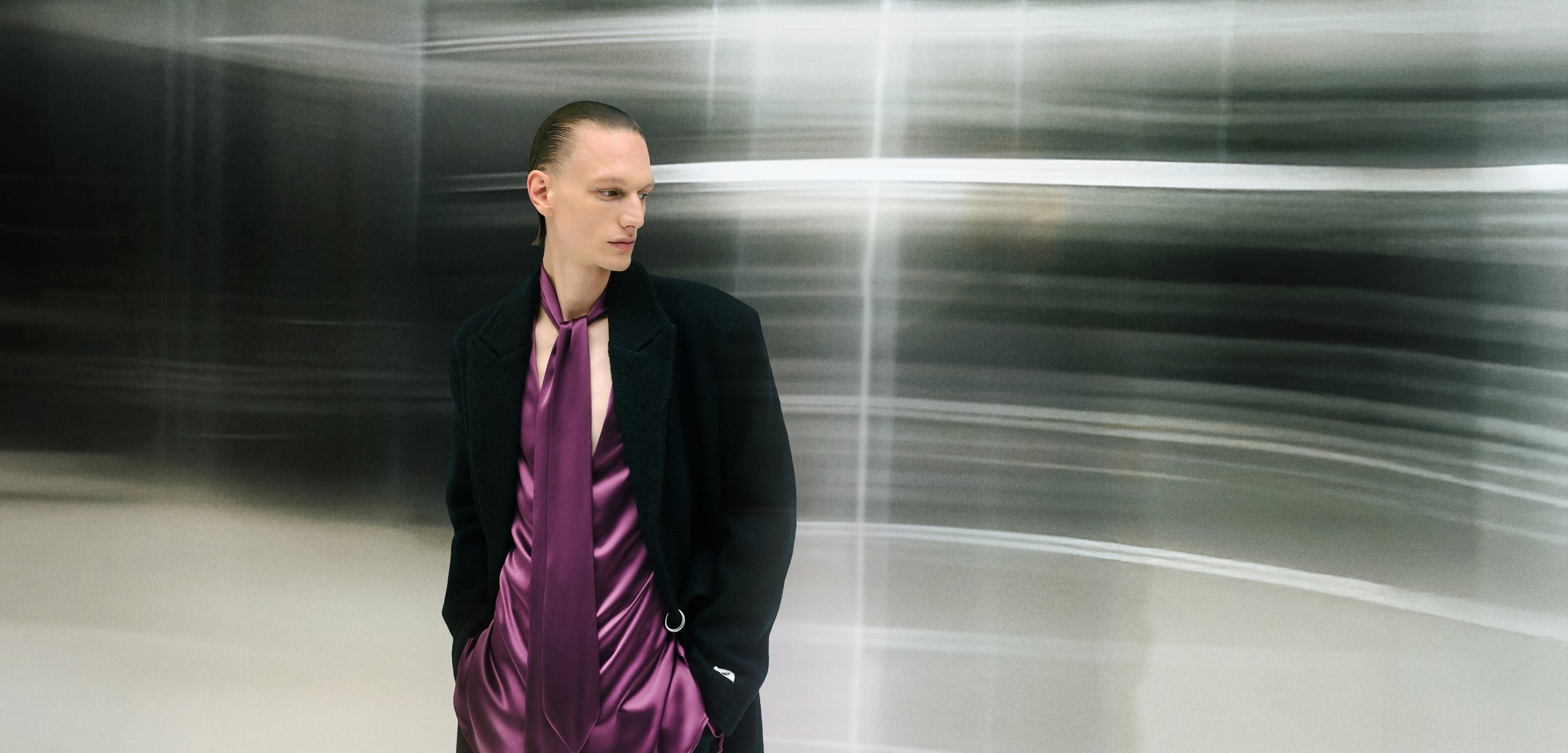 Model wearing a black coat over a purple satin shirt and tie. The background is blurred with soft, smooth lines and a metallic sheen.
