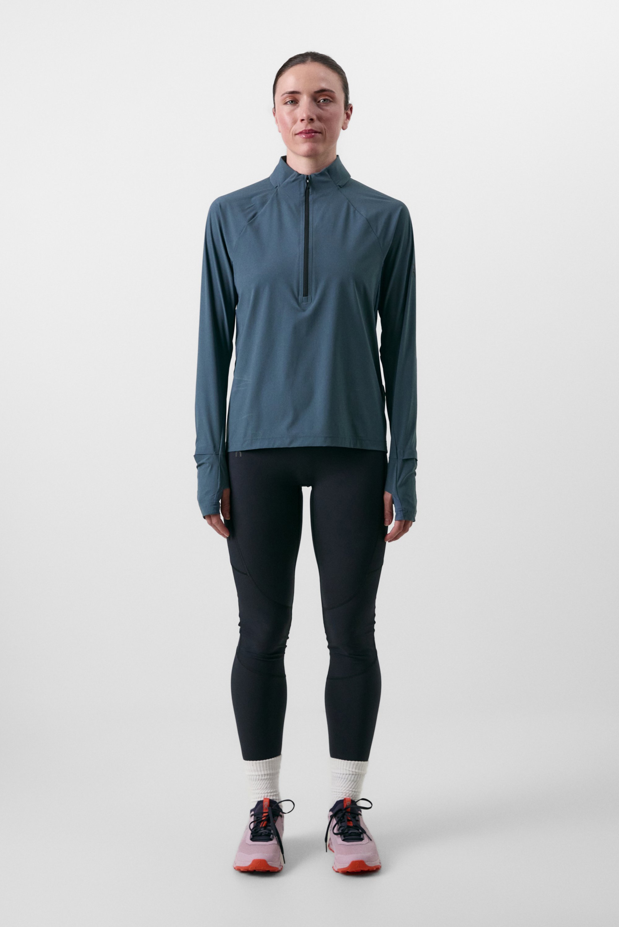 Athletic woman wearing a zip-up teal top, black leggings, and running shoes, standing against a plain white background.