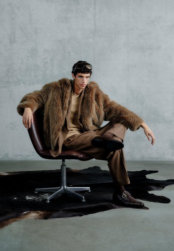 Bărbat tânăr în pardesiu maro de blană și costum, șezând cu un picior peste celălalt pe un scaun rotativ, deasupra unui covor întunecat din piele de animal, în fața unui perete de beton.