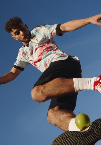 Man in een bloemenvoetbalshirt en zwarte short trapt een groene appel in de lucht tegen een heldere blauwe lucht.
