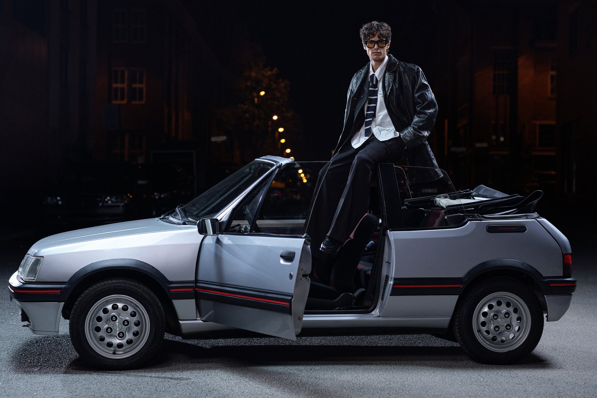 Una persona con estilo se encuentra al borde de un coche convertible vintage, vestida con una chaqueta de cuero, camisa y corbata, en una calle poco iluminada.