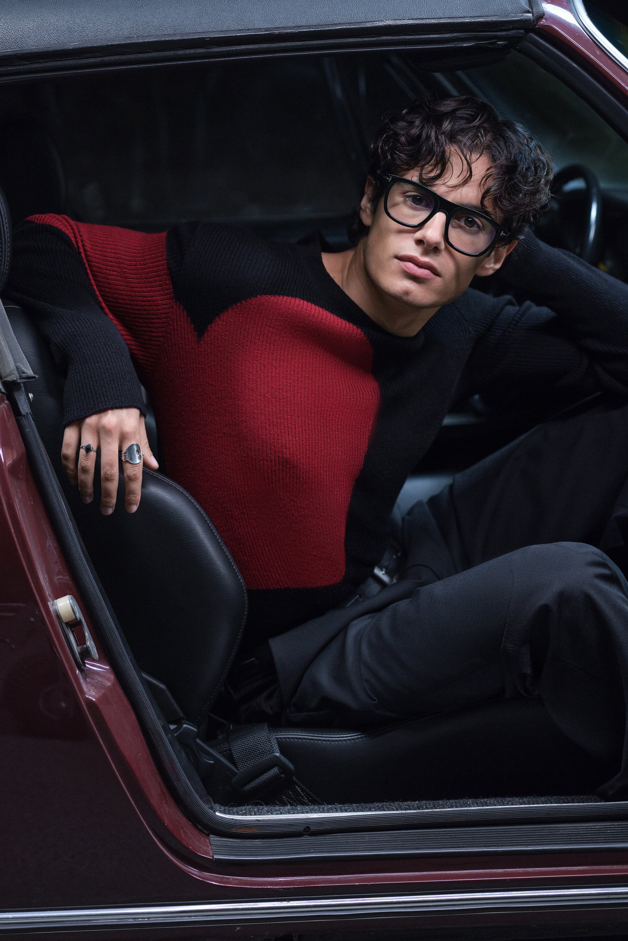 Una persona joven con cabello rizado se sienta en la puerta abierta de un coche, vistiendo un suéter negro y rojo y gafas, mostrando anillos de moda en su mano.