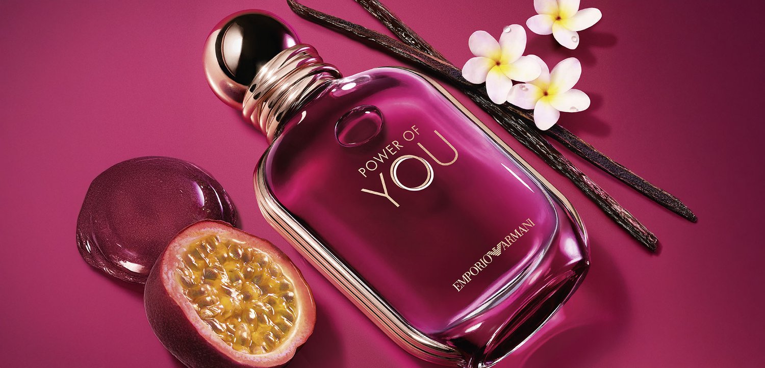 Lila Emporio Armani "Power of You" Parfümflasche mit halbierten Passionsfrüchten, Vanilleschoten und weißen Blumen auf rosa Hintergrund.