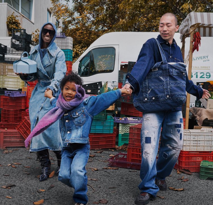 Three individuals are holding hands in a market setting, dressed in denim attire. One of them is carrying a light blue container, and they are surrounded by vibrant crates.