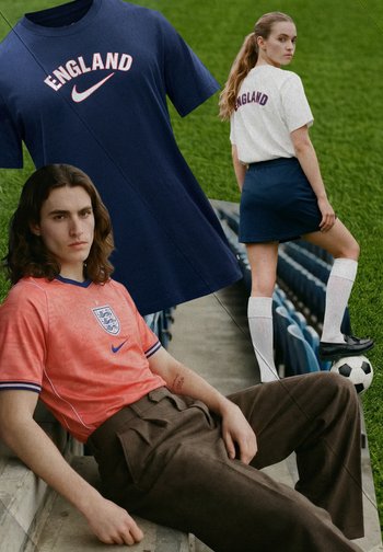 Junger Mann im orangefarbenen England-Fußballtrikot lehnt sich auf den Stadionstufen zurück; Frau steht auf dem Spielfeld mit dem Fuß auf dem Ball, trägt ein England-T-Shirt und einen Rock.