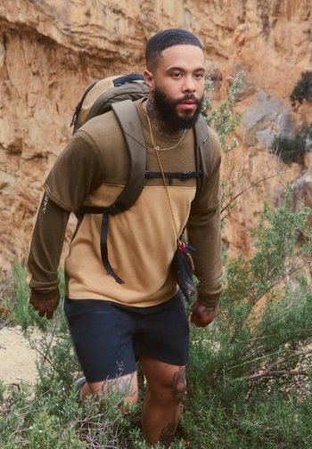 Man with a backpack and tattoos hiking uphill through rocky terrain with green shrubs, wearing a beige and brown long-sleeve shirt and black shorts.