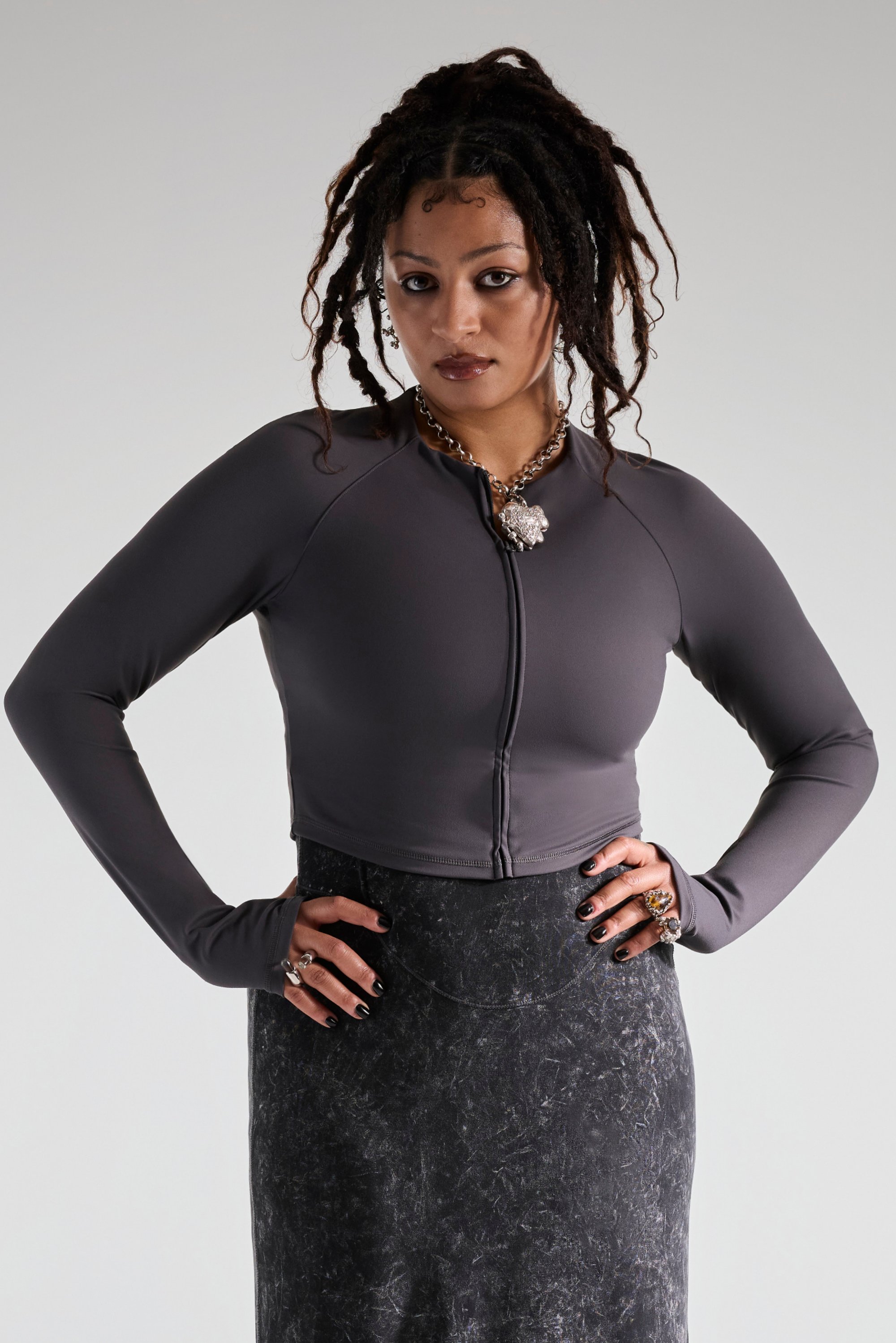 Una persona con rastas hasta los hombros posa con confianza, vistiendo una blusa gris corta y una larga falda negra, mostrando joyas ornamentadas.