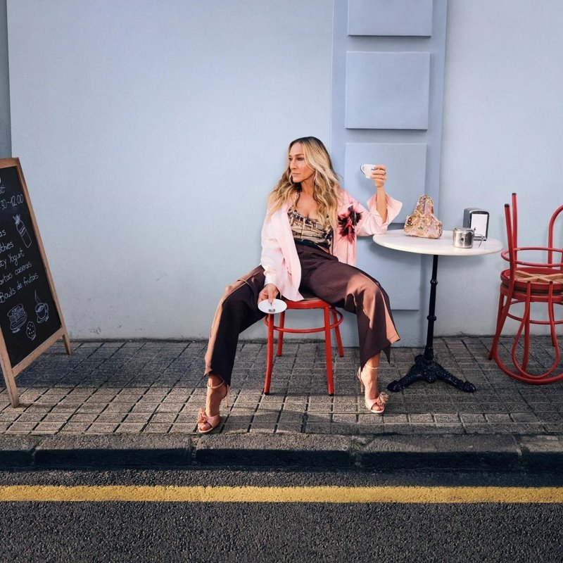 Een vrouw zit op een rode stoel, houdt een kopje vast, voor een café met een tafel, een krijtbordmenu en een beige handtas in de buurt.