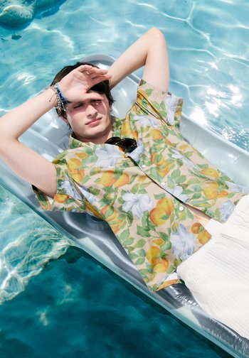 Tânăr cu cămașă cu flori și pantaloni albi culcat pe o plută de piscină, ferindu-și ochii de soare cu o mână.