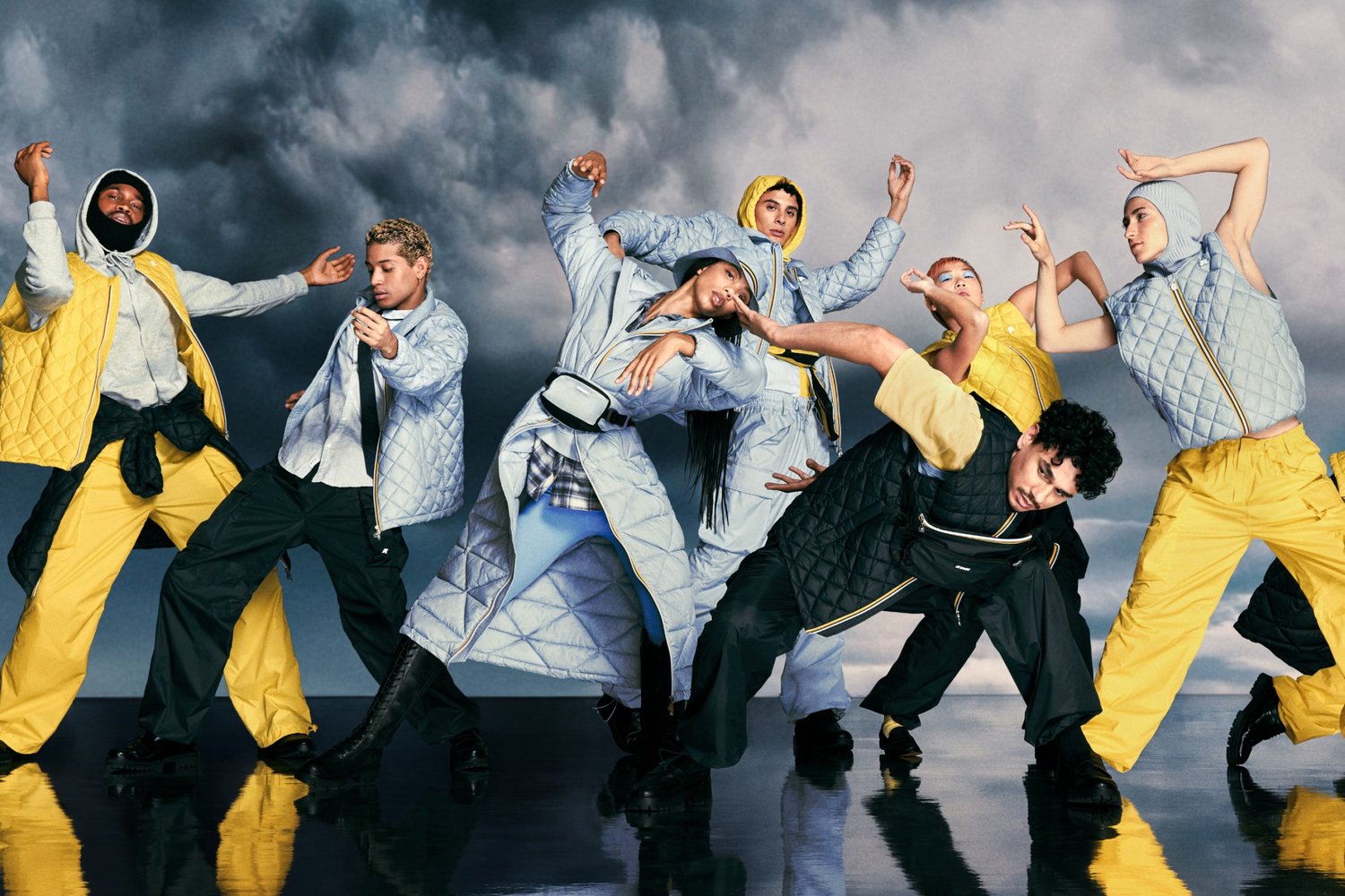 Nove persone vestite in modo elegante, con outfit vivaci, ballano espressivamente su uno sfondo drammatico e nuvoloso, mostrando movimento e stile.